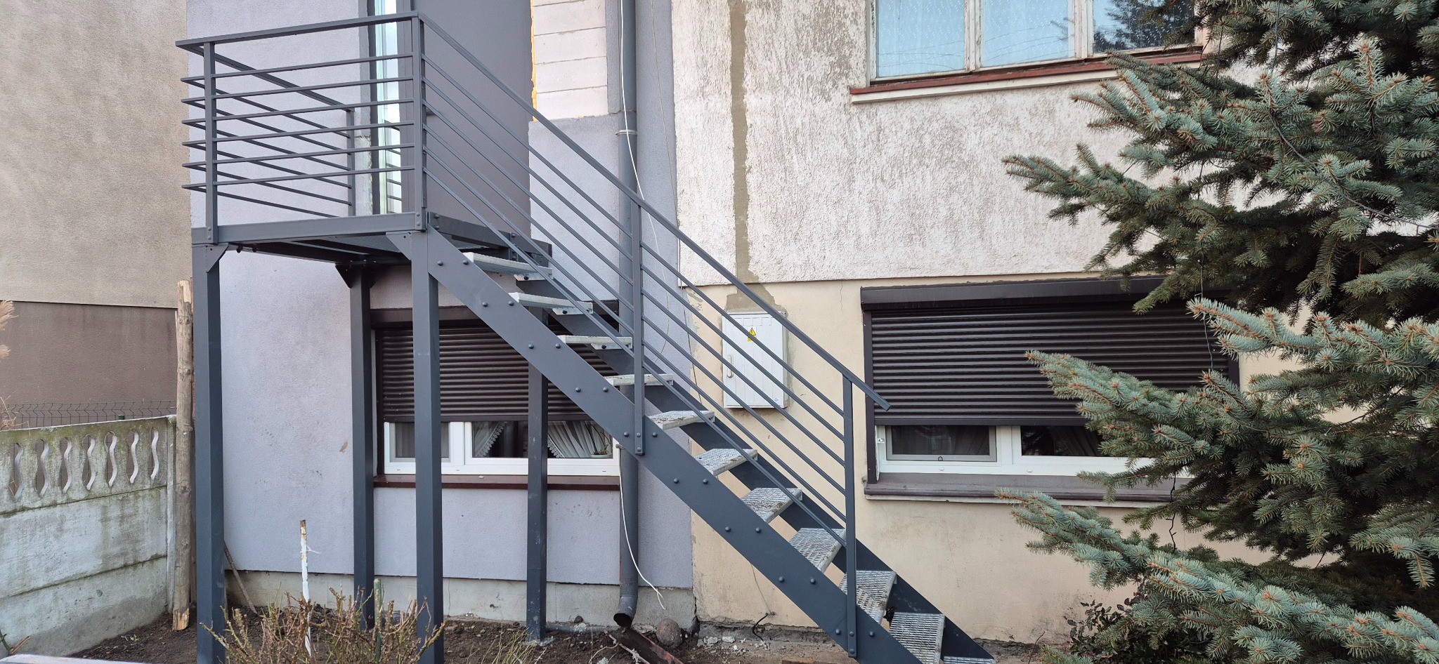 Zewnętrzne schody metalowe z balustradą, prowadzące do drzwi na piętrze, pomalowane na szaro, z widocznymi nitowanymi stopniami i prostą konstrukcją.