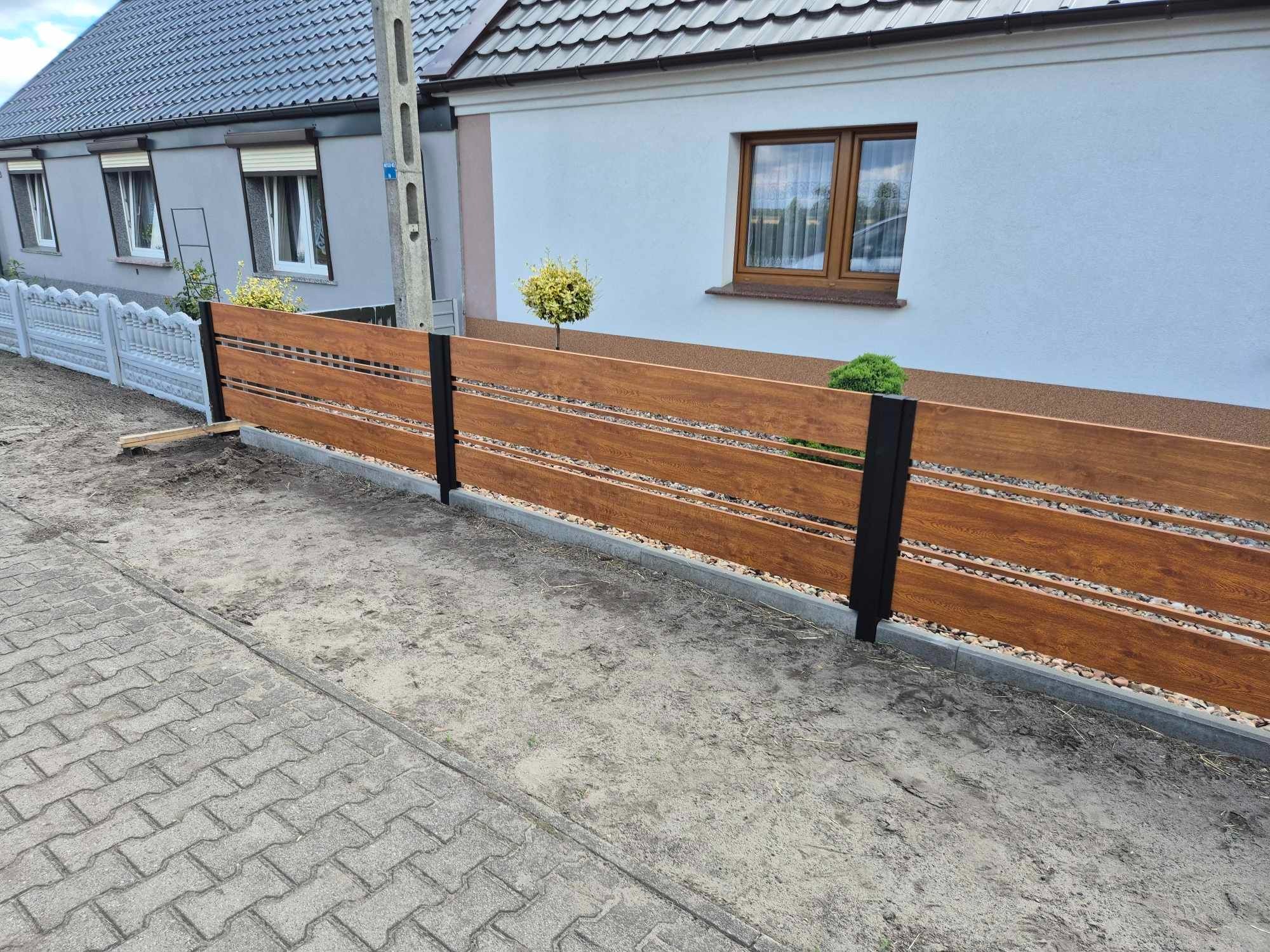 Nowoczesne ogrodzenie panelowe z poziomych desek w kolorze drewna, z czarnymi metalowymi słupkami, oddzielające posesję od chodnika z kostki brukowej.