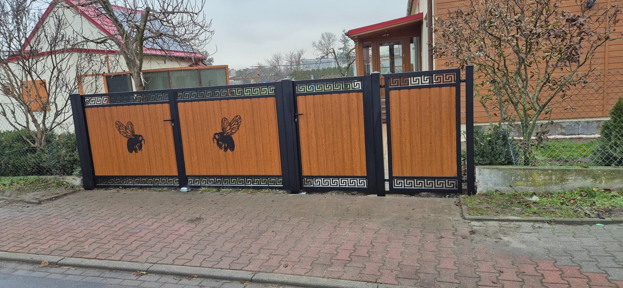 Nowoczesna brama wjazdowa z drewnopodobnym wypełnieniem i czarną ramą, ozdobiona motywem pszczoły i greckim wzorem, na tle domu jednorodzinnego.