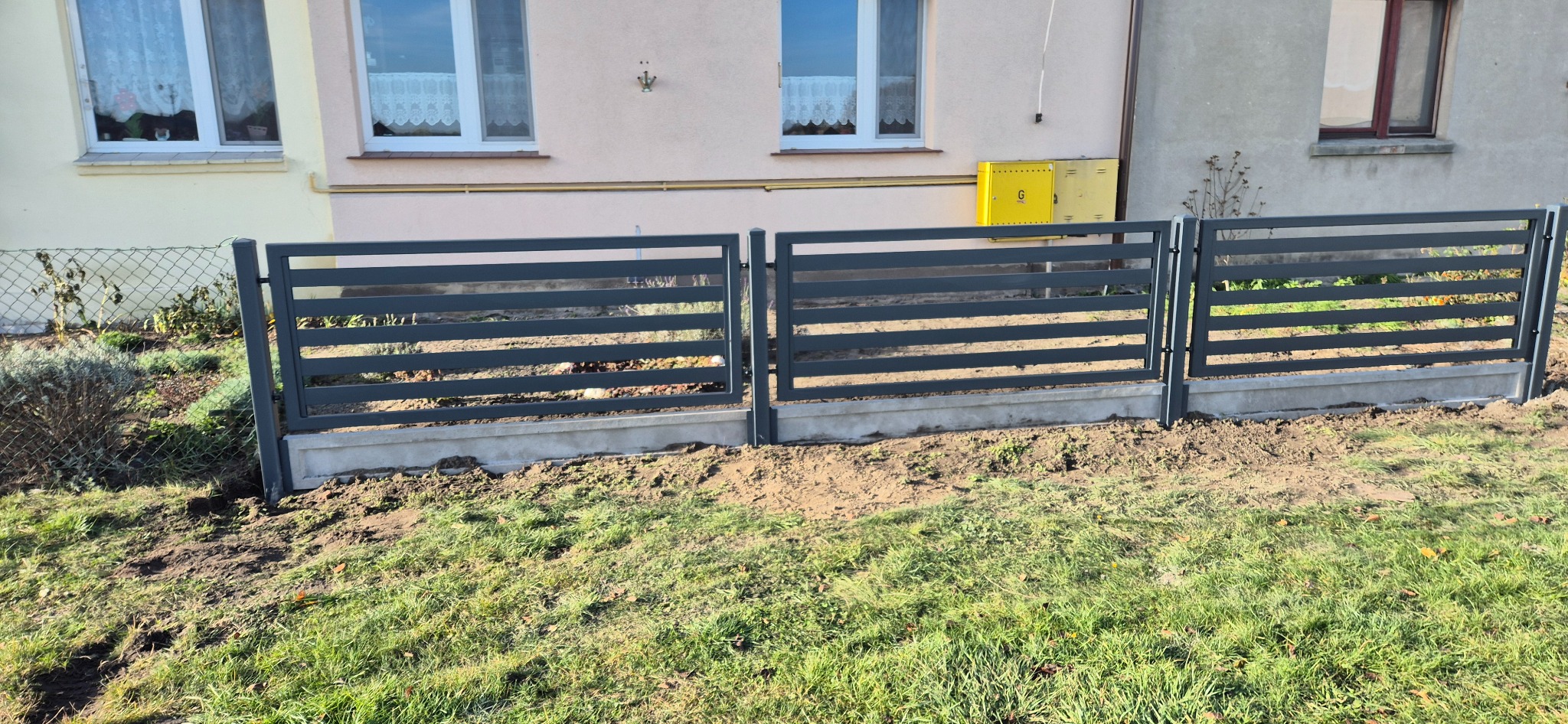 Nowoczesne, poziome przęsła metalowe, szare, zamontowane na betonowej podmurówce, widoczne świeże ślady po montażu w postaci ziemi i trawy, w tle budynek mieszkalny z żółtą skrzynką gazową.