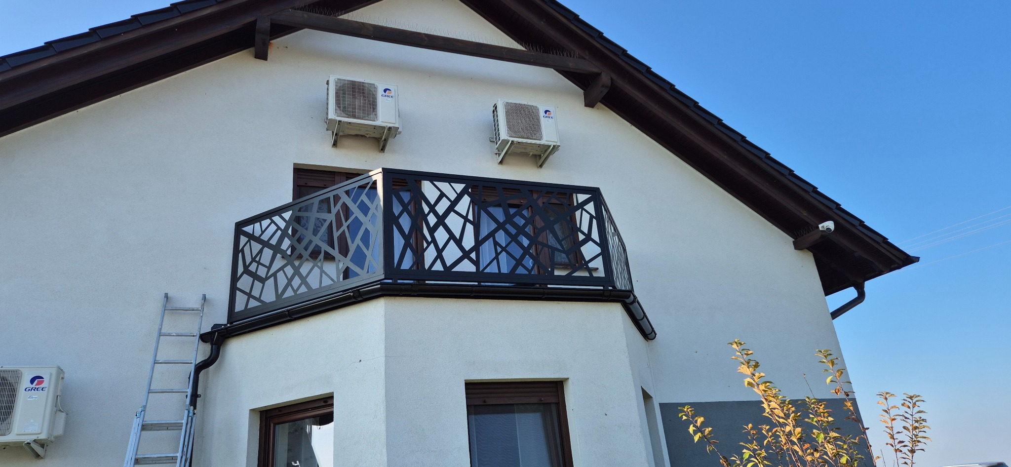 Czarna, ażurowa balustrada balkonowa o geometrycznym wzorze, zamontowana na białym budynku z dwoma klimatyzatorami i drabiną opartą o ścianę.