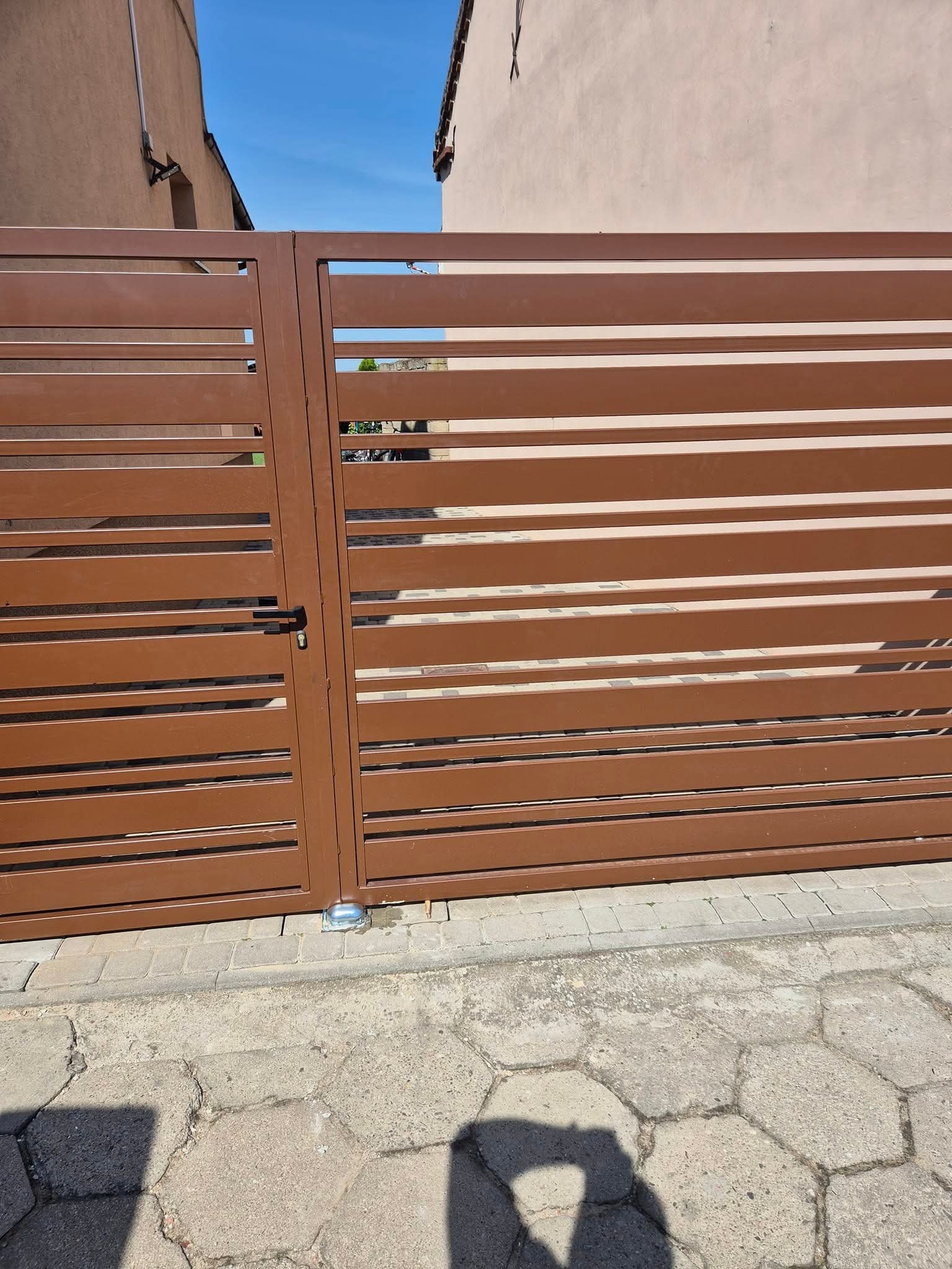 Nowoczesna, brązowa brama wjazdowa z poziomymi panelami, zamontowana na posesji z kostką brukową. Widoczny zamek i zawiasy. Brama w stylu minimalistycznym.