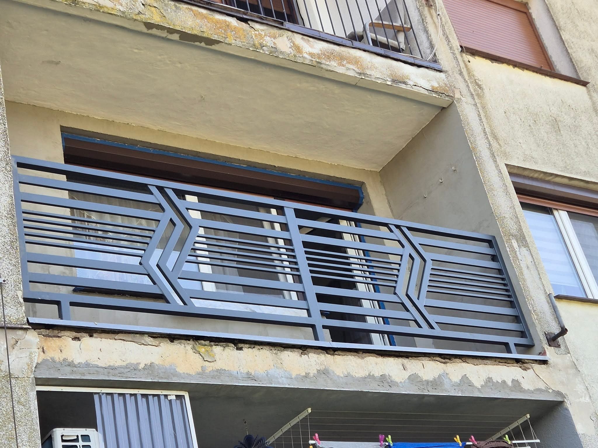 Szara, metalowa balustrada balkonowa z geometrycznym wzorem na tle elewacji budynku. Widoczne detale mocowania i otoczenie balkonu.