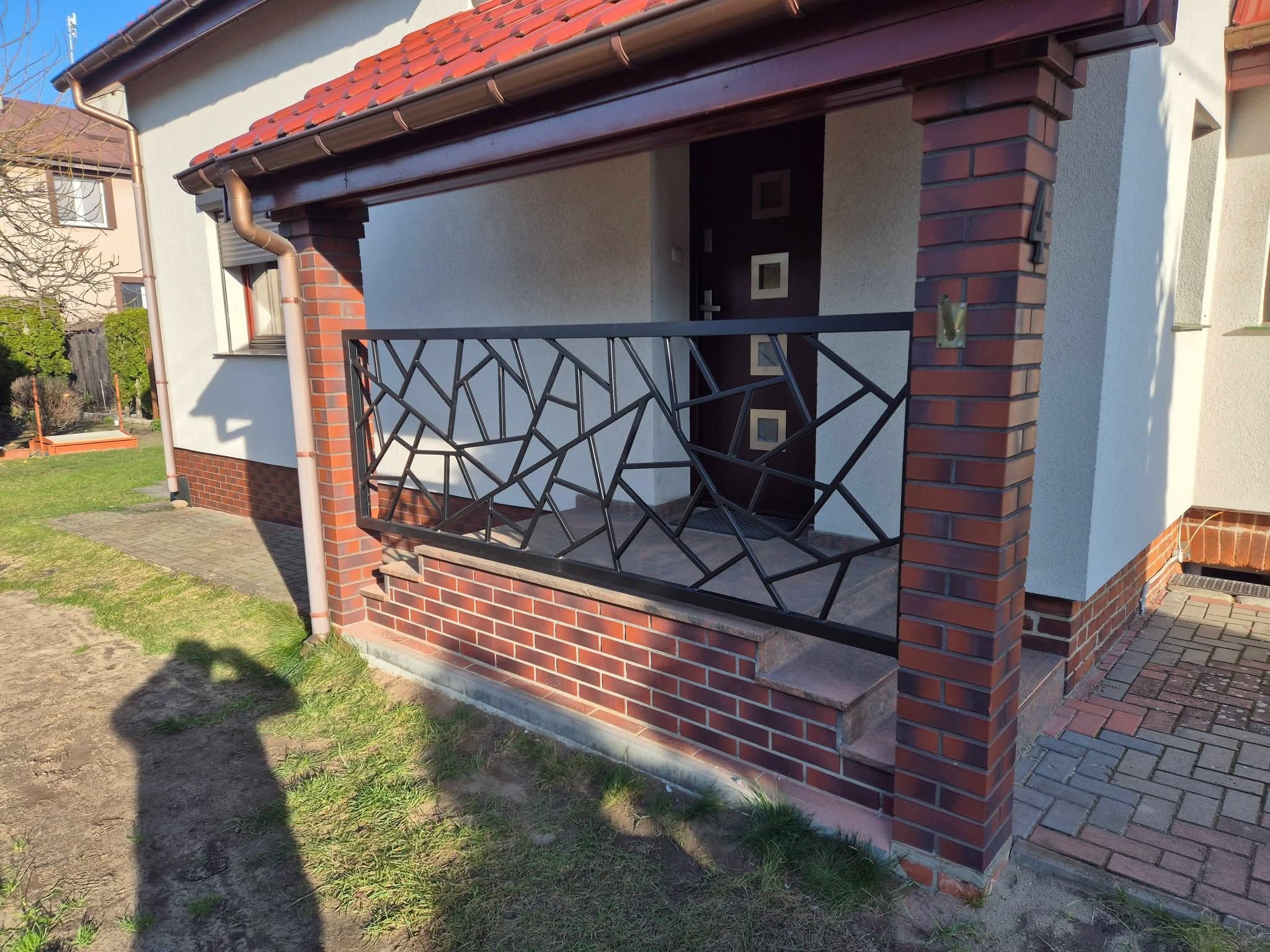 Nowoczesna, czarna balustrada metalowa o geometrycznym wzorze, zamontowana na ceglanym podeście przed wejściem do domu. Widoczny cień osoby robiącej zdjęcie.