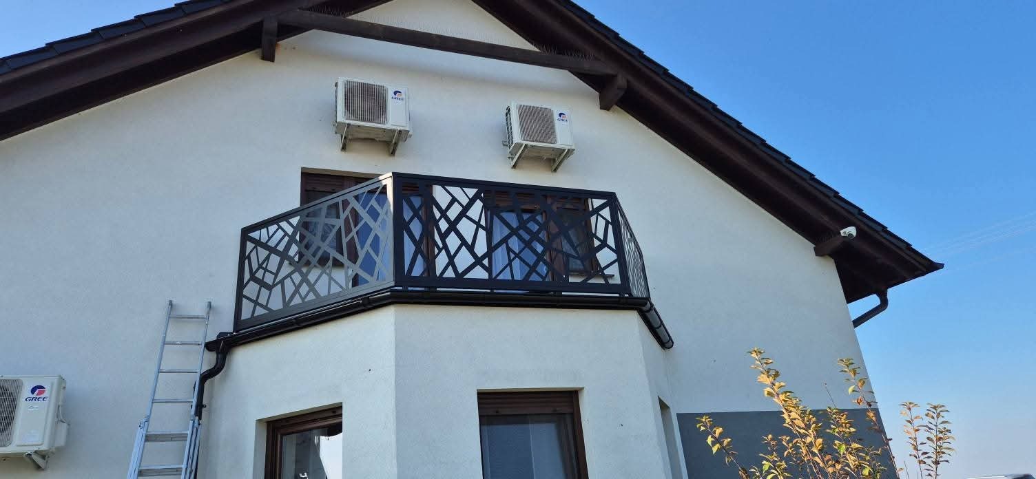 Czarna, metalowa balustrada balkonowa z geometrycznym wzorem na tle białej elewacji domu w Raszewach. Widoczna drabina i klimatyzatory.