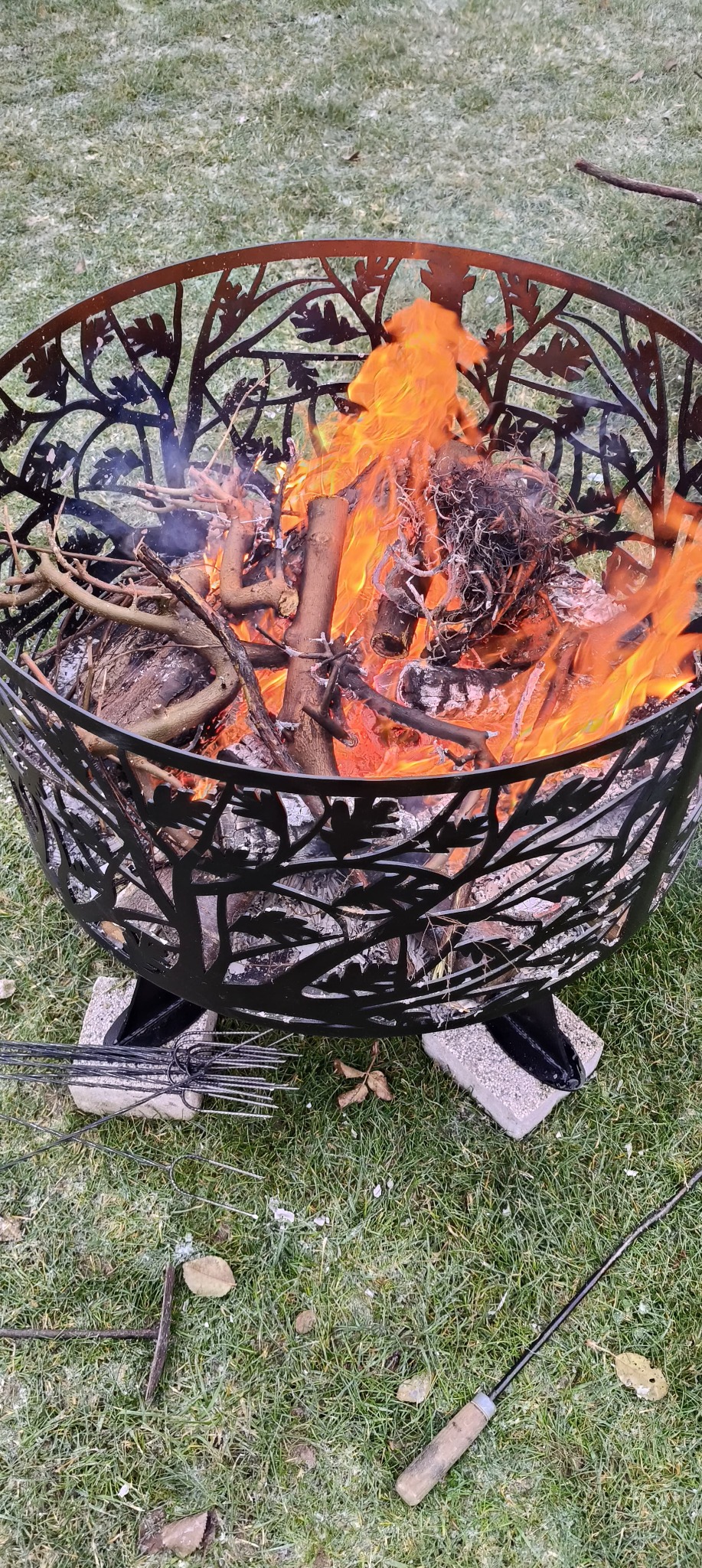 Metalowy kosz paleniskowy z wyciętym motywem drzewa, wypełniony płonącym drewnem, stoi na trawie. Widoczne narzędzia do ogniska.