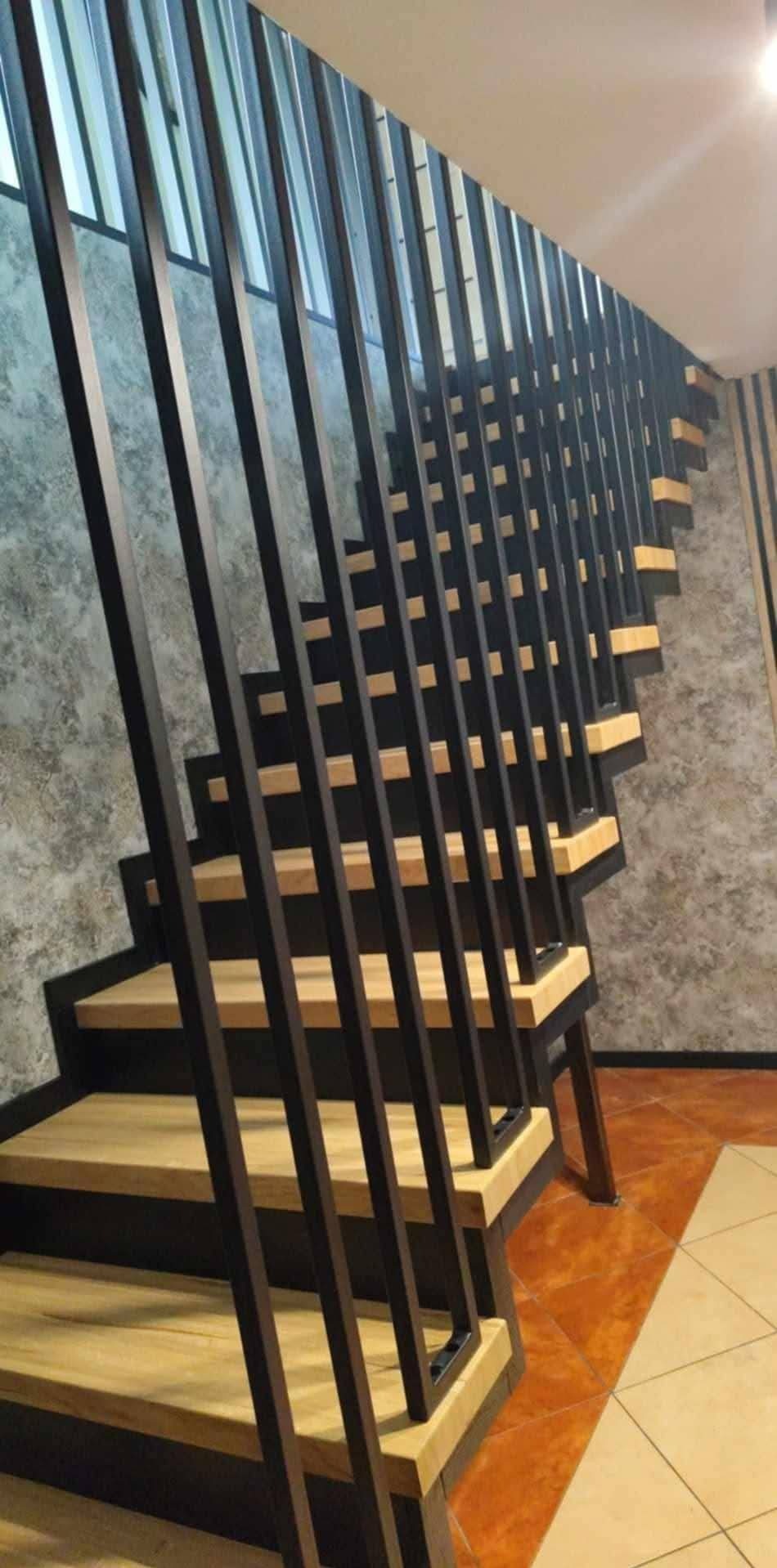 Nowoczesne schody z drewnianymi stopniami i minimalistyczną, metalową balustradą z pionowych prętów. Widok z dołu, podkreślający geometrię konstrukcji.