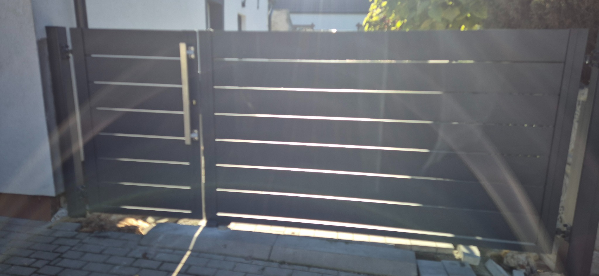 Nowoczesna, metalowa brama wjazdowa w kolorze grafitowym z poziomymi szczeblami, widoczna z bliska, z delikatnym efektem światła słonecznego.