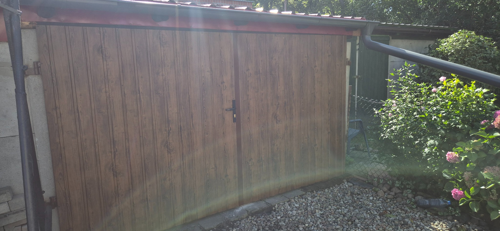 Dwuskrzydłowa brama garażowa z brązowymi panelami imitującymi drewno, z czarną klamką na tle zieleni i szarej kostki brukowej.