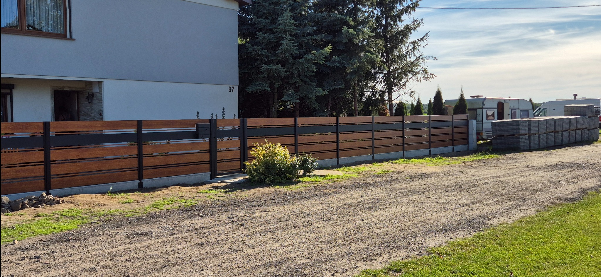 Nowoczesne ogrodzenie posesji z drewnianymi panelami i czarnymi, metalowymi słupkami na tle domu z numerem 97 i zieleni. Widoczny fragment drogi gruntowej.