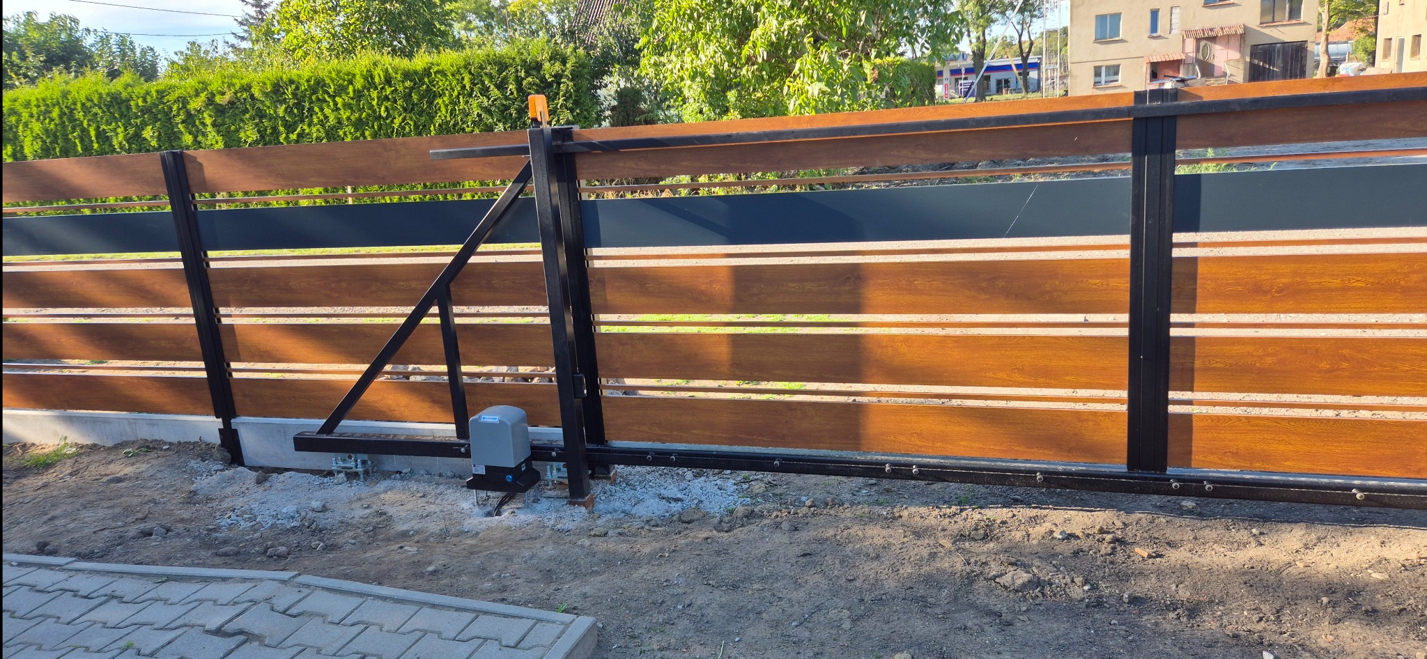 Nowoczesna brama przesuwna z drewnopodobnymi panelami i czarną ramą, widoczny napęd elektryczny. Brama zamontowana, otoczenie w trakcie prac wykończeniowych.