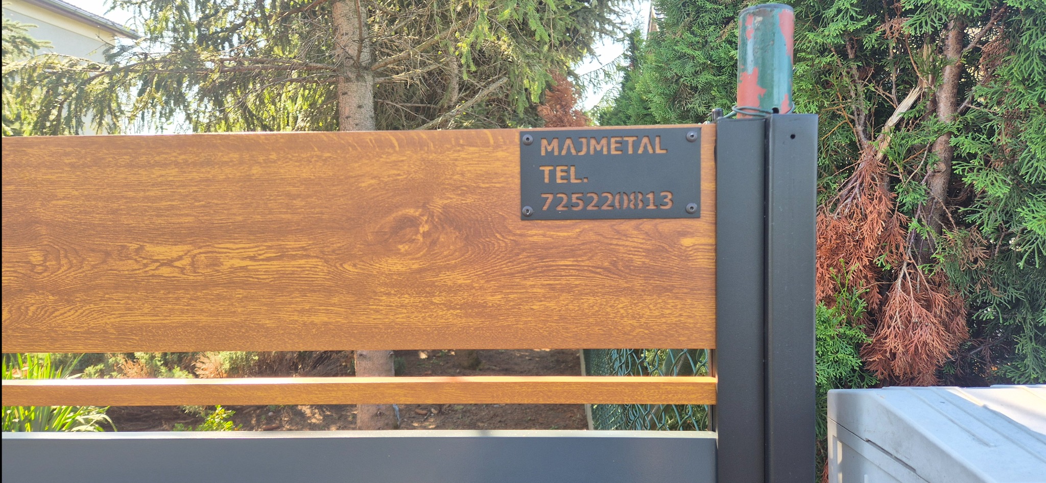 Fragment bramy wjazdowej z drewnopodobnym wypełnieniem i tabliczką z nazwą firmy MAJMETAL oraz numerem telefonu 725220813, na tle zieleni.