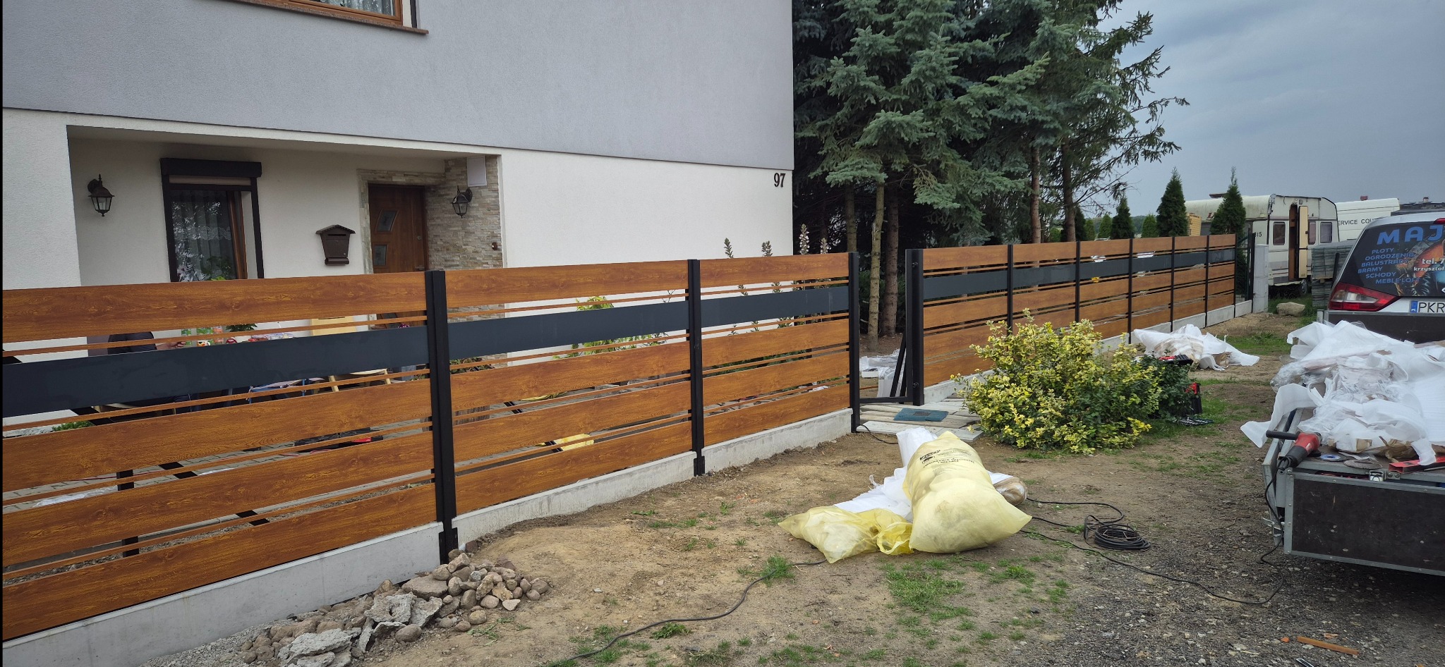 Nowoczesne ogrodzenie panelowe z drewnopodobnymi przęsłami i czarnymi słupkami na tle domu z numerem 97, widoczne worki z odpadami po montażu.
