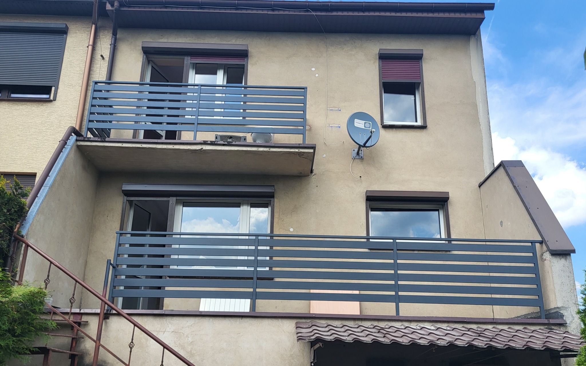 Szare, poziome balustrady balkonowe zamontowane na dwukondygnacyjnym budynku z beżową elewacją, widoczne schody wejściowe z metalu i antena satelitarna na ścianie.