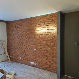 Usługi budowlane NEW HOME Kamil Richert - Ściana z cegły klinkierowej z oświetleniem LED, widoczne przygotowania do prac wykończeniowych, zabezpieczona folią podłoga, drabina i narzędzia malarskie.