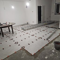 Usługi budowlane NEW HOME Kamil Richert - Układanie szarych płytek podłogowych z użyciem pomarańczowych klipsów poziomujących w przestronnym, jasnym pomieszczeniu; widoczna drabina aluminiowa i okrągły stół z narzędziami w tle.