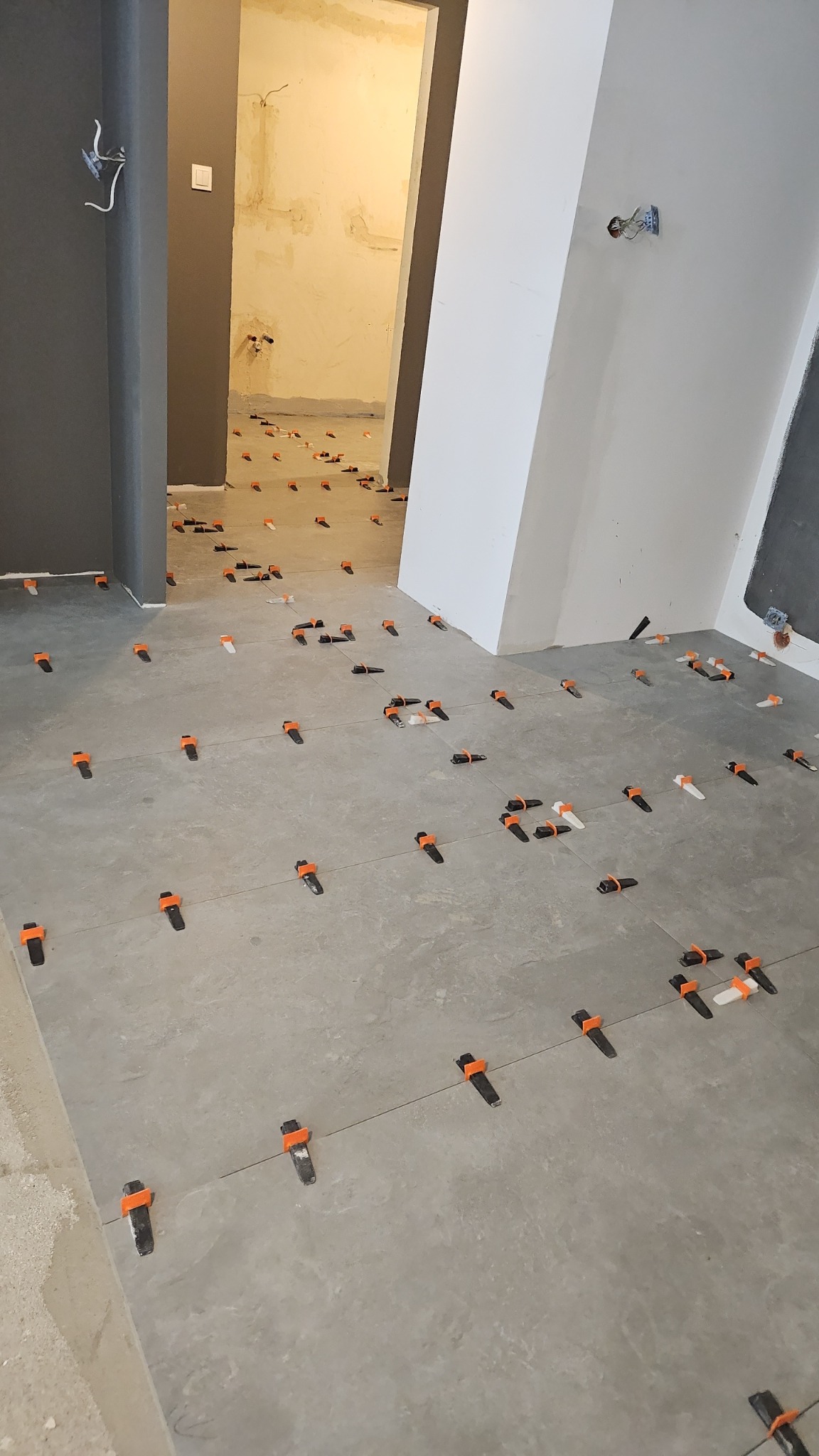 Układanie szarych płytek podłogowych z użyciem pomarańczowych klipsów poziomujących, widoczne niedokończone ściany w tle.