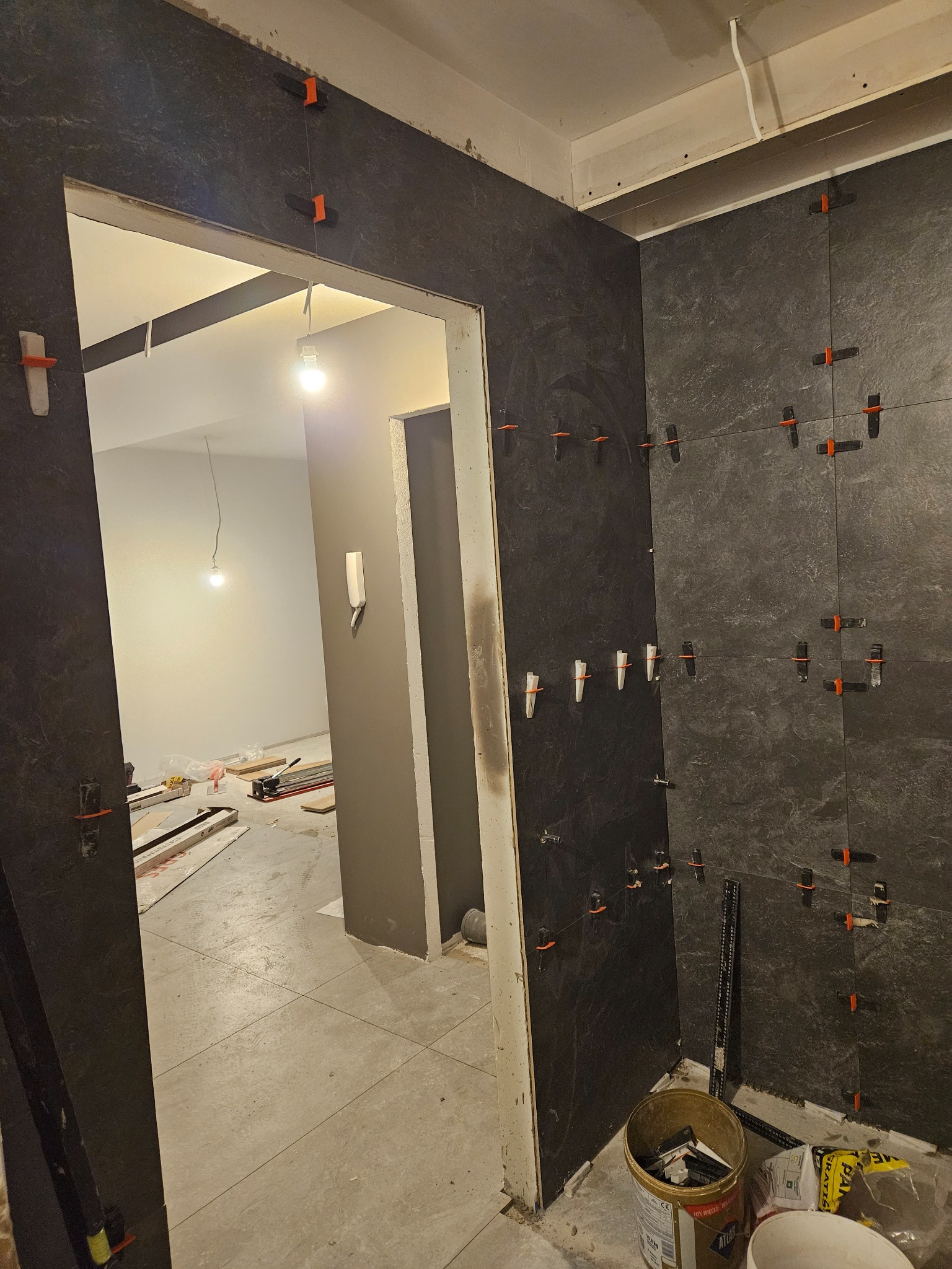 Montaż ciemnych płytek na ścianie w łazience, z użyciem klipsów poziomujących; widoczny fragment podłogi i sąsiedniego pomieszczenia w trakcie prac wykończeniowych.