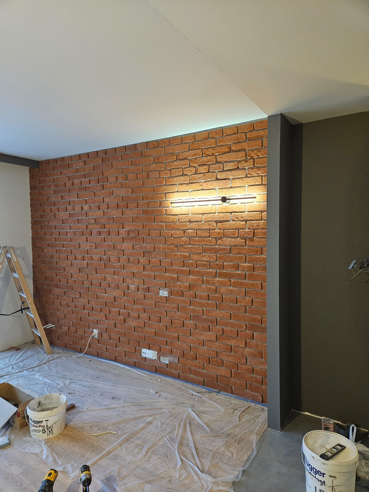 Ściana z cegły klinkierowej z oświetleniem LED, widoczne przygotowania do prac wykończeniowych, zabezpieczona folią podłoga, drabina i narzędzia malarskie.