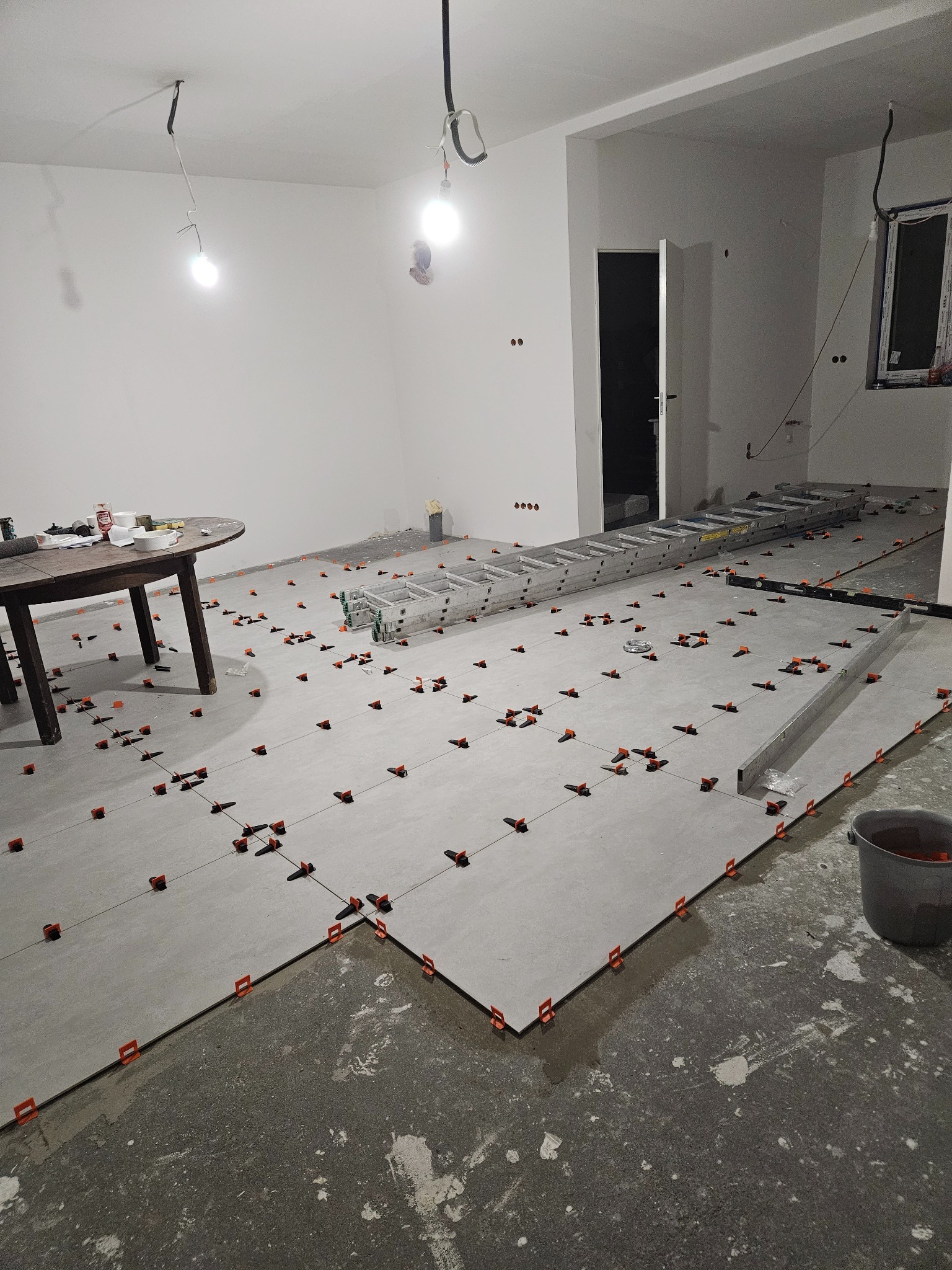 Układanie szarych płytek podłogowych z użyciem pomarańczowych klipsów poziomujących w przestronnym, jasnym pomieszczeniu; widoczna drabina aluminiowa i okrągły stół z narzędziami w tle.