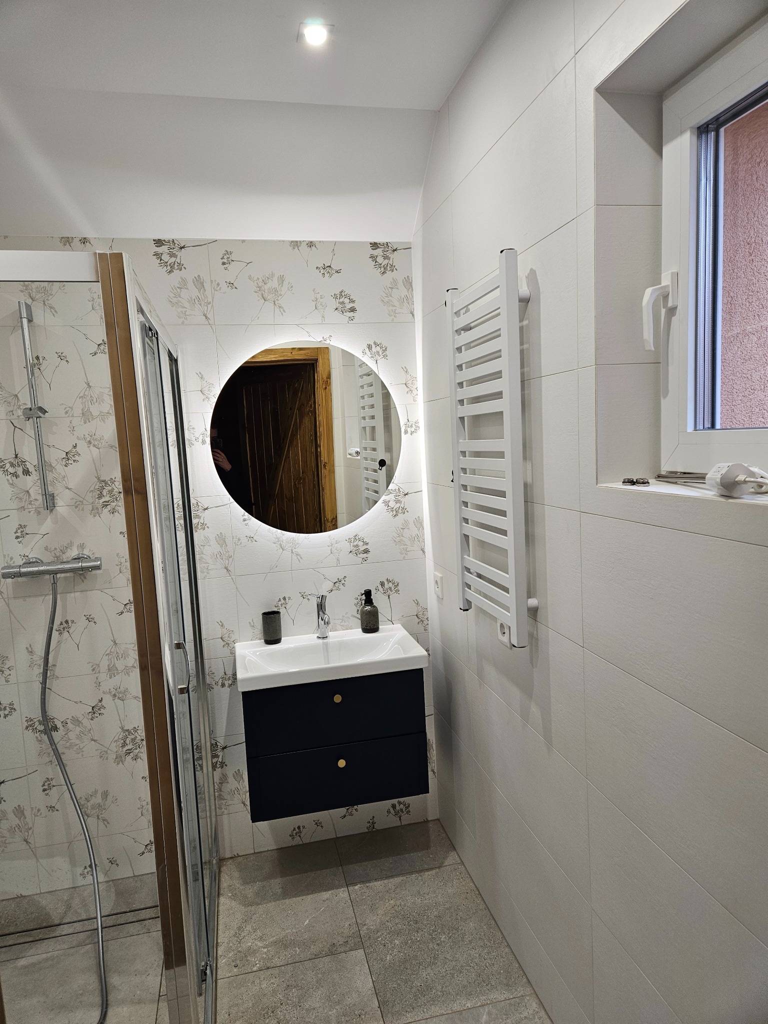 Nowoczesna łazienka z kabiną prysznicową, umywalką z szafką w kolorze granatowym z okrągłym lustrem podświetlanym LED i grzejnikiem drabinkowym w kolorze białym, wykończona płytkami w dwóch wzorach...