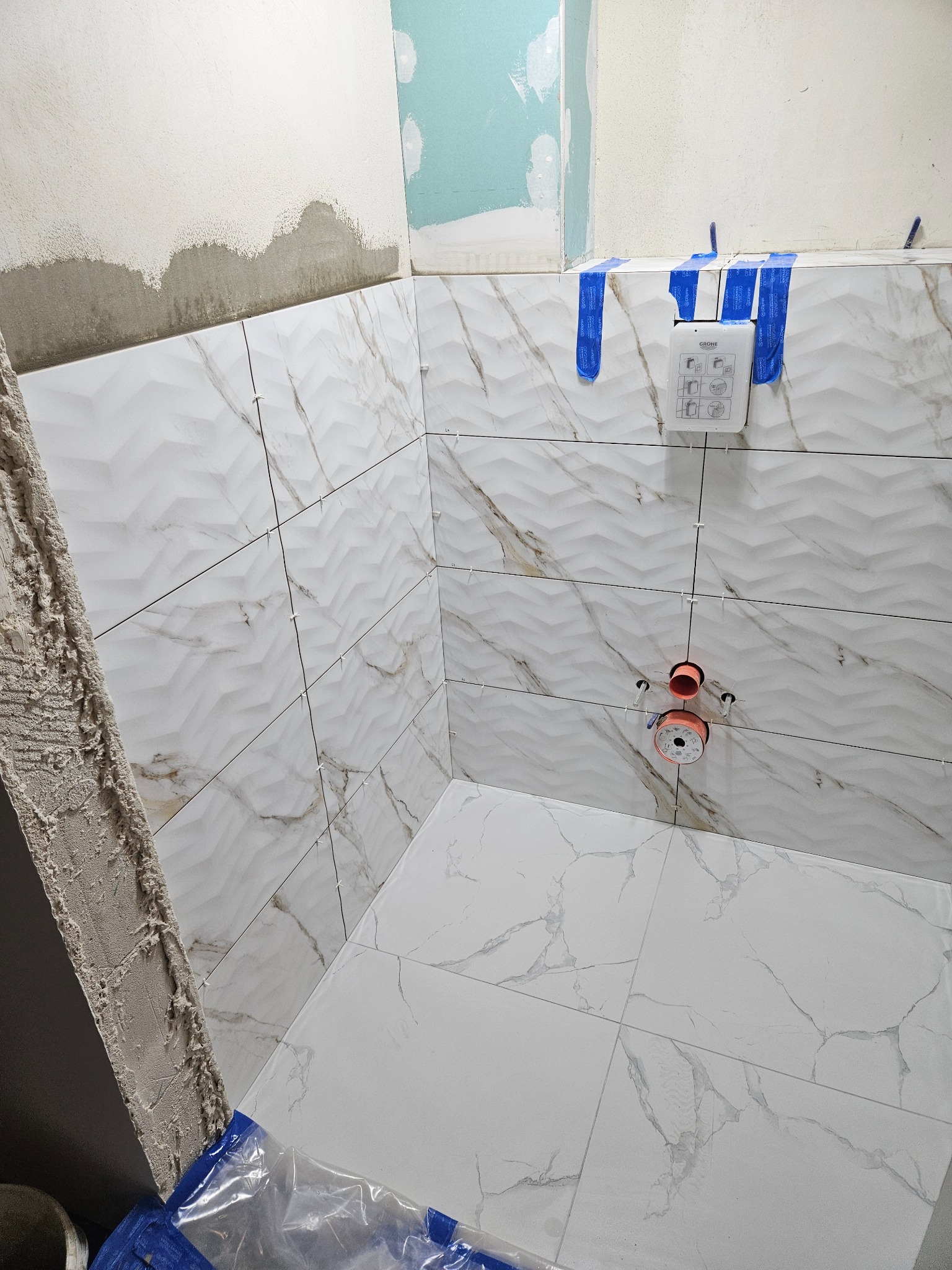 Świeżo ułożone, białe płytki z marmurkowym wzorem na ścianach i podłodze w łazience, widoczne elementy instalacji wodnej i nieotynkowany fragment ściany.
