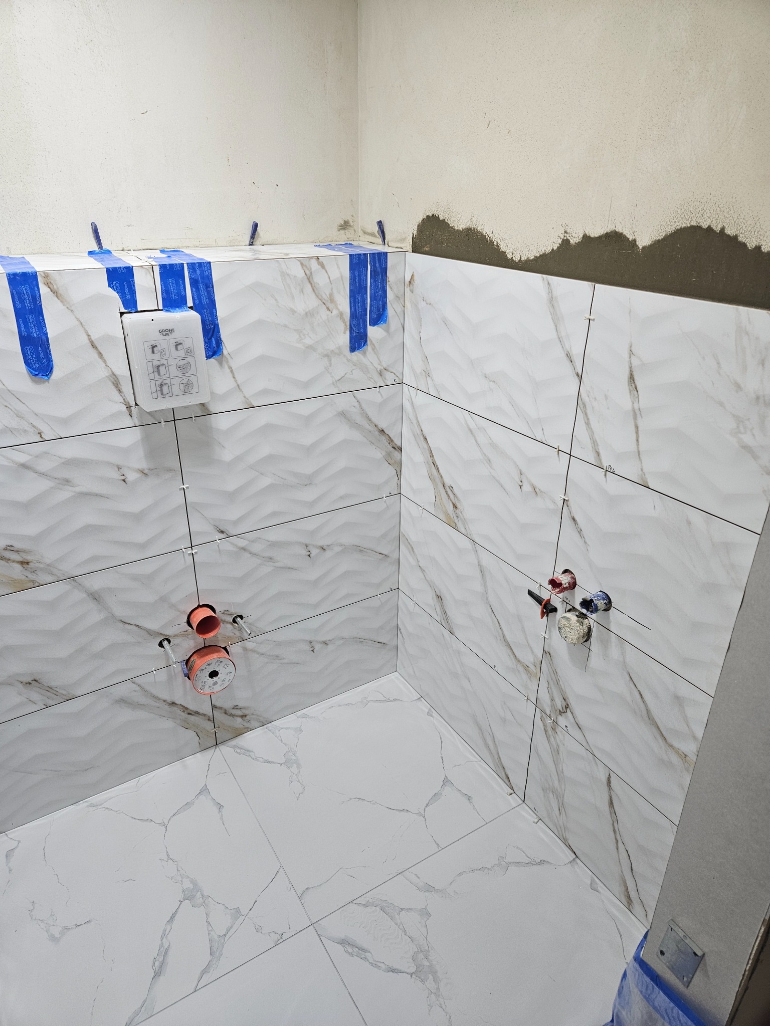 Białe płytki imitujące marmur, ułożone na ścianach i podłodze łazienki w trakcie montażu, zabezpieczone niebieską taśmą, widoczne elementy instalacji wodnej i stelaż podtynkowy WC.