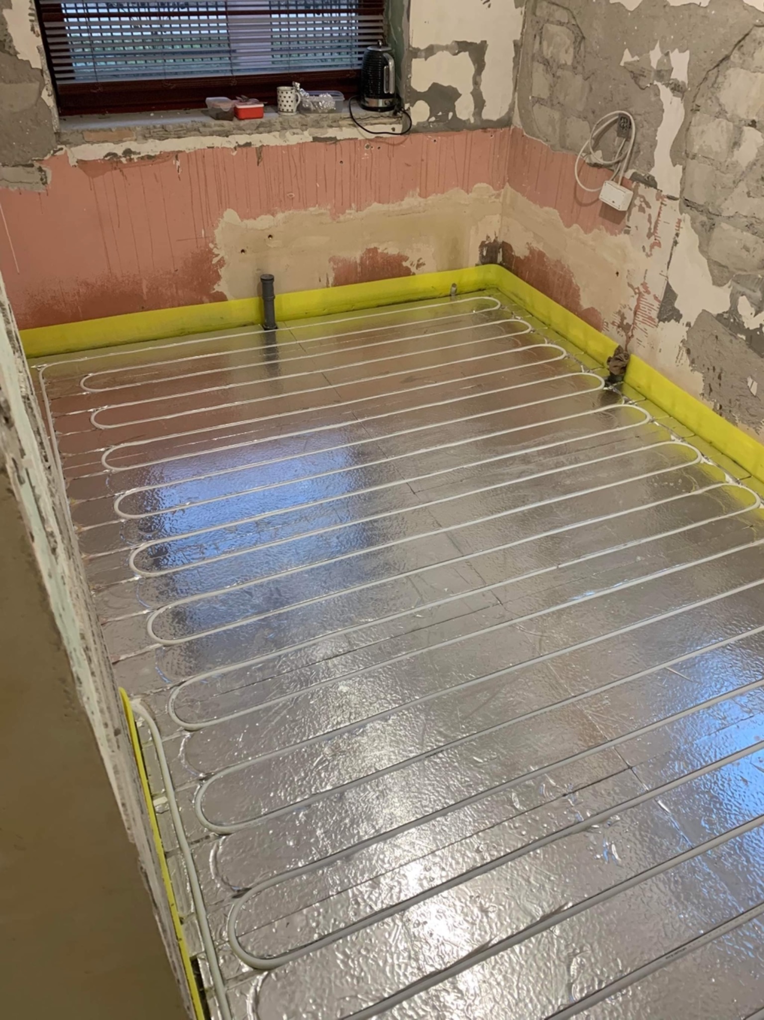 Szare rury ogrzewania podłogowego ułożone na srebrnej folii izolacyjnej w surowym pomieszczeniu z częściowo otynkowanymi ścianami i oknem w tle.