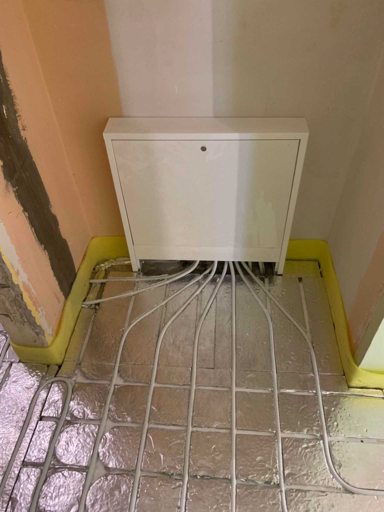 Rozłożona instalacja ogrzewania podłogowego na folii aluminiowej, z widoczną białą skrzynką rozdzielczą i przewodami wychodzącymi z niej, przy ścianach zabezpieczonych żółtą pianką dylatacyjną.