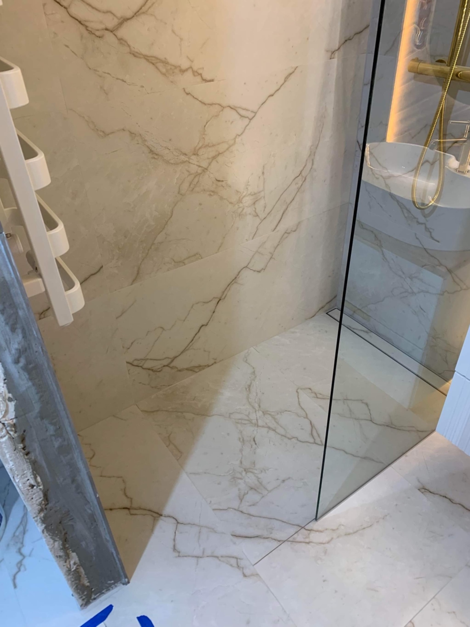 Wykończona kabina prysznicowa z marmurowymi płytkami, widoczny odpływ liniowy i złota armatura.