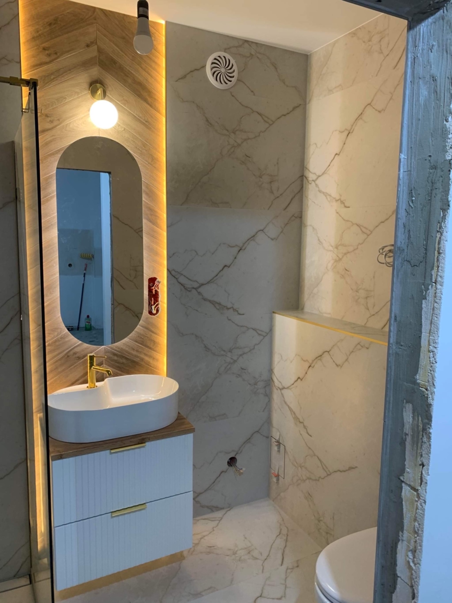 Wykończona łazienka z marmurowymi płytkami, podświetlanym lustrem owalnym, białą umywalką na szafce z złotymi uchwytami i złotą armaturą, widok na toaletę i surowy beton w otworze drzwiowym.