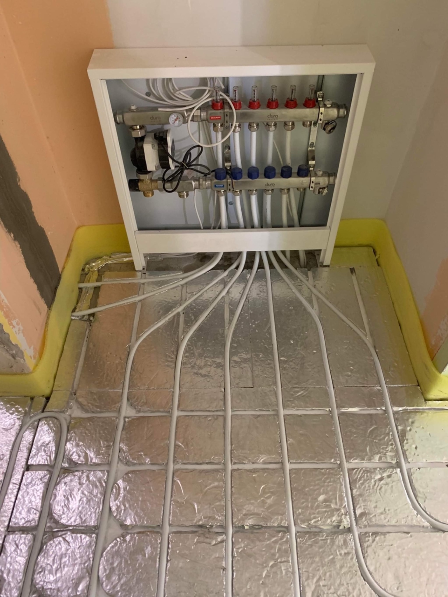 Rozdzielacz ogrzewania podłogowego Duro w białej obudowie, rury rozprowadzone na folii izolacyjnej z żółtą pianką przy ścianach, w trakcie montażu.