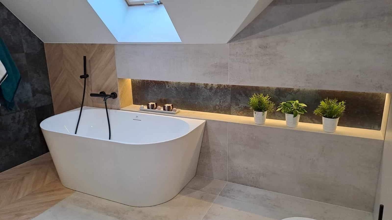 Nowoczesna łazienka na poddaszu z wolnostojącą białą wanną, czarną armaturą, szarą glazurą i podświetlaną wnęką z roślinami doniczkowymi.