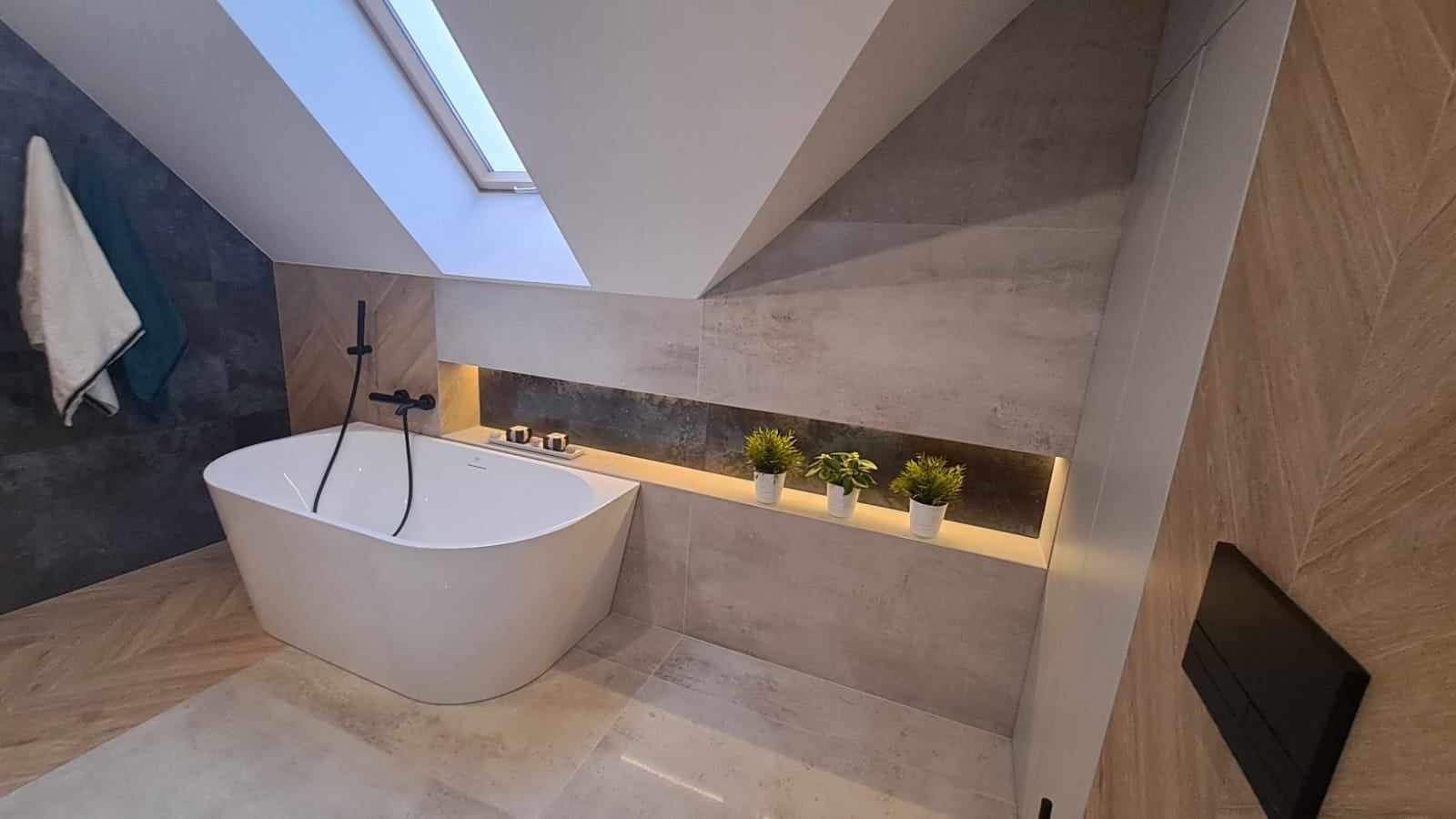 Nowoczesna łazienka na poddaszu z wolnostojącą białą wanną, czarną armaturą, szarą glazurą, oknem dachowym i dekoracyjną półką z podświetleniem i trzema małymi roślinami.