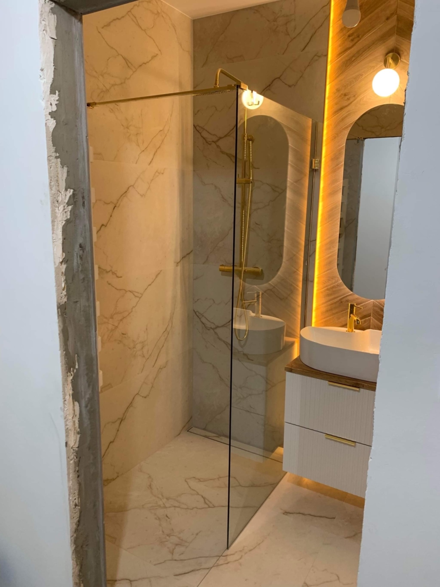 Wnętrze łazienki w trakcie wykańczania: widoczna kabina prysznicowa z elementami w kolorze złota, umywalka nablatowa na szafce z podświetleniem LED, marmurowe płytki na ścianach i podłodze, surowy...