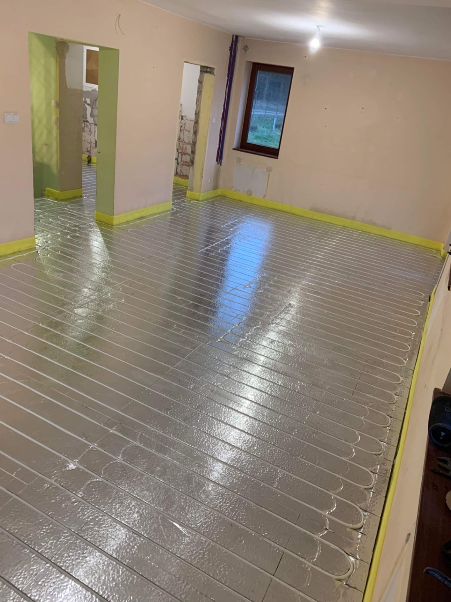 Rozłożona instalacja ogrzewania podłogowego na folii aluminiowej w trakcie remontu, widoczne przygotowane przejścia w ścianach i okno.