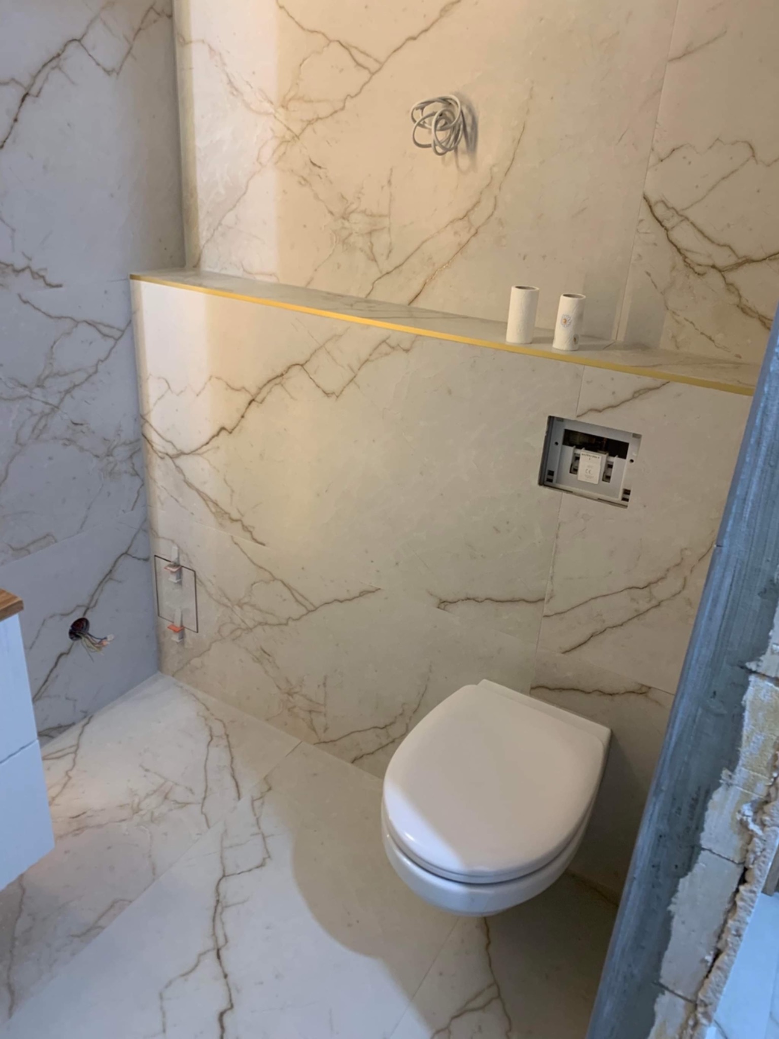 Wnętrze łazienki w trakcie wykończenia, widoczne marmurowe płytki na ścianach i podłodze, podtynkowa spłuczka, wisząca toaleta oraz wystające przewody elektryczne.