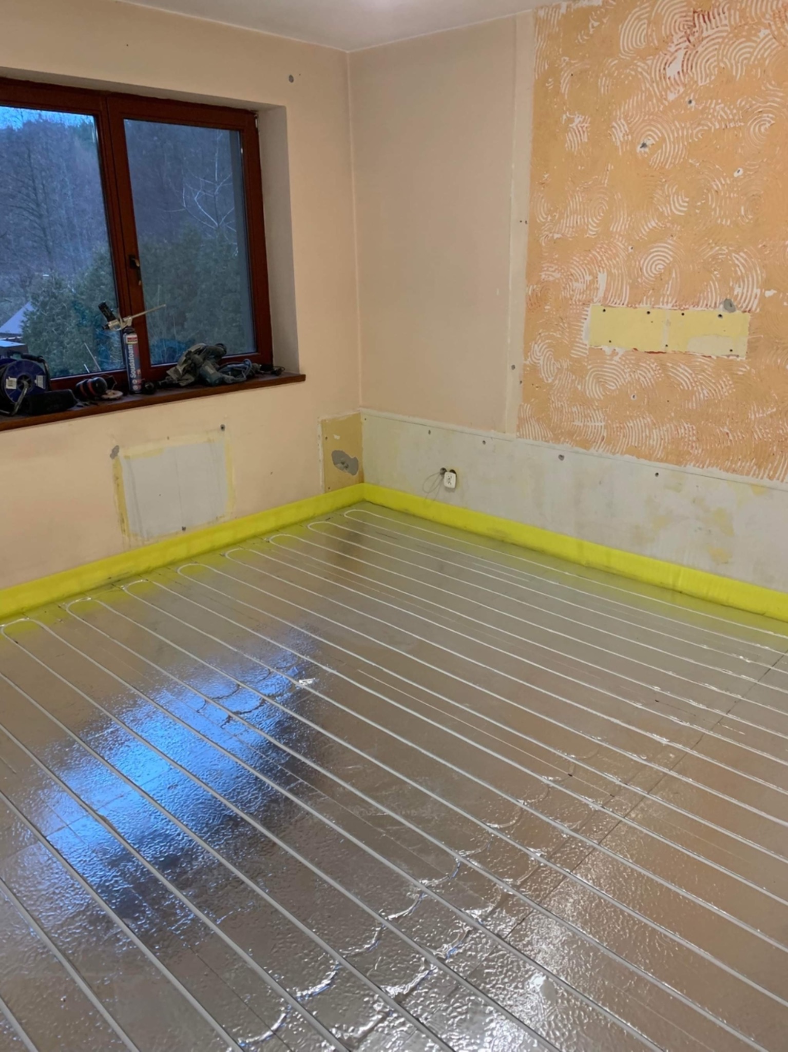 Instalacja ogrzewania podłogowego z folią aluminiową w pomieszczeniu w Mszanowie, widoczne przygotowane ściany do dalszych prac wykończeniowych i okno z widokiem na zewnątrz.