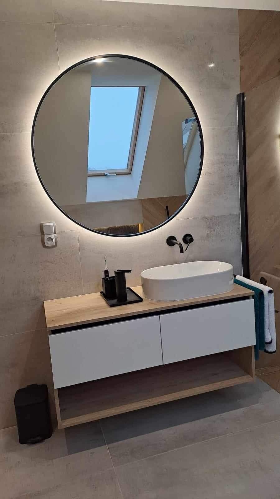 Nowoczesna łazienka z owalną umywalką nablatową, okrągłym lustrem z podświetleniem LED i widokiem na okno dachowe, utrzymana w stonowanej kolorystyce.