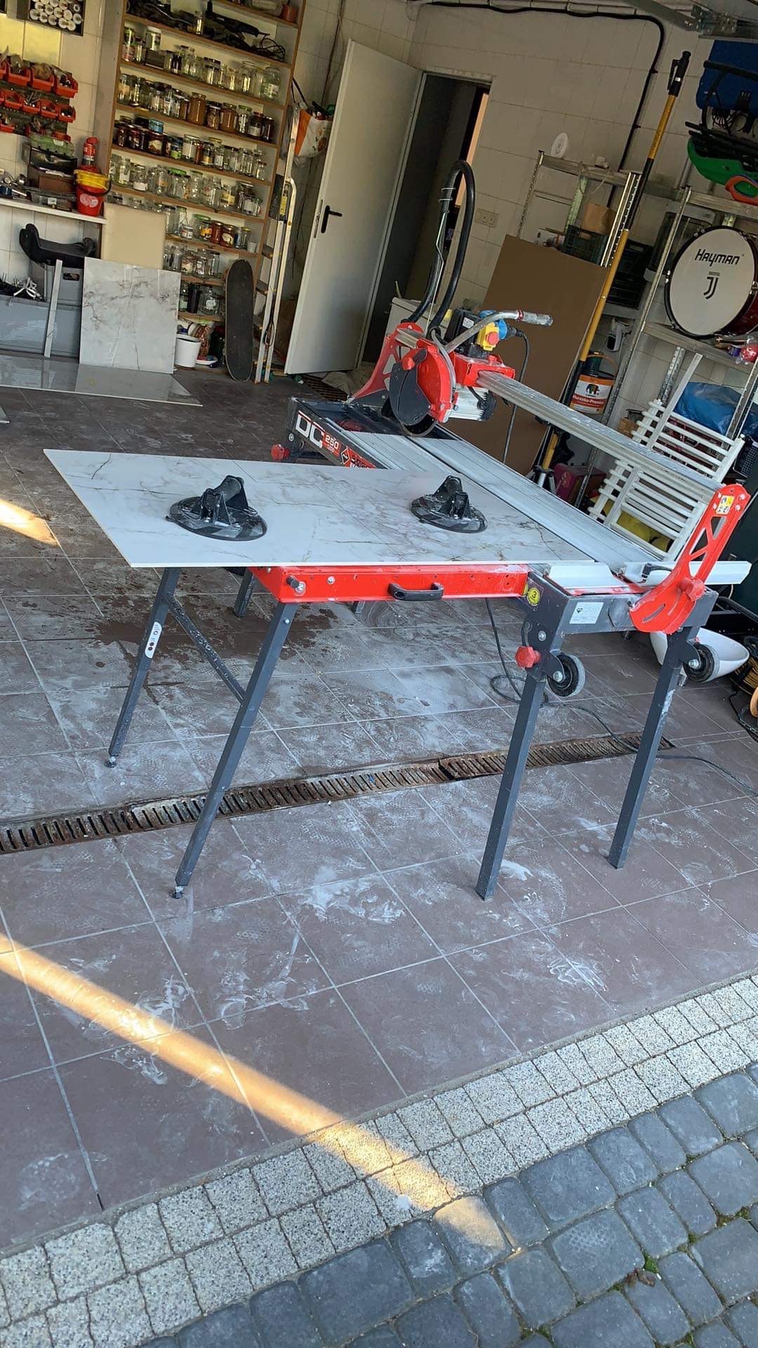 Elektryczna przecinarka do płytek z marmurową płytą na stole roboczym, widoczne przyssawki do mocowania, tło warsztatowe z półkami i narzędziami.