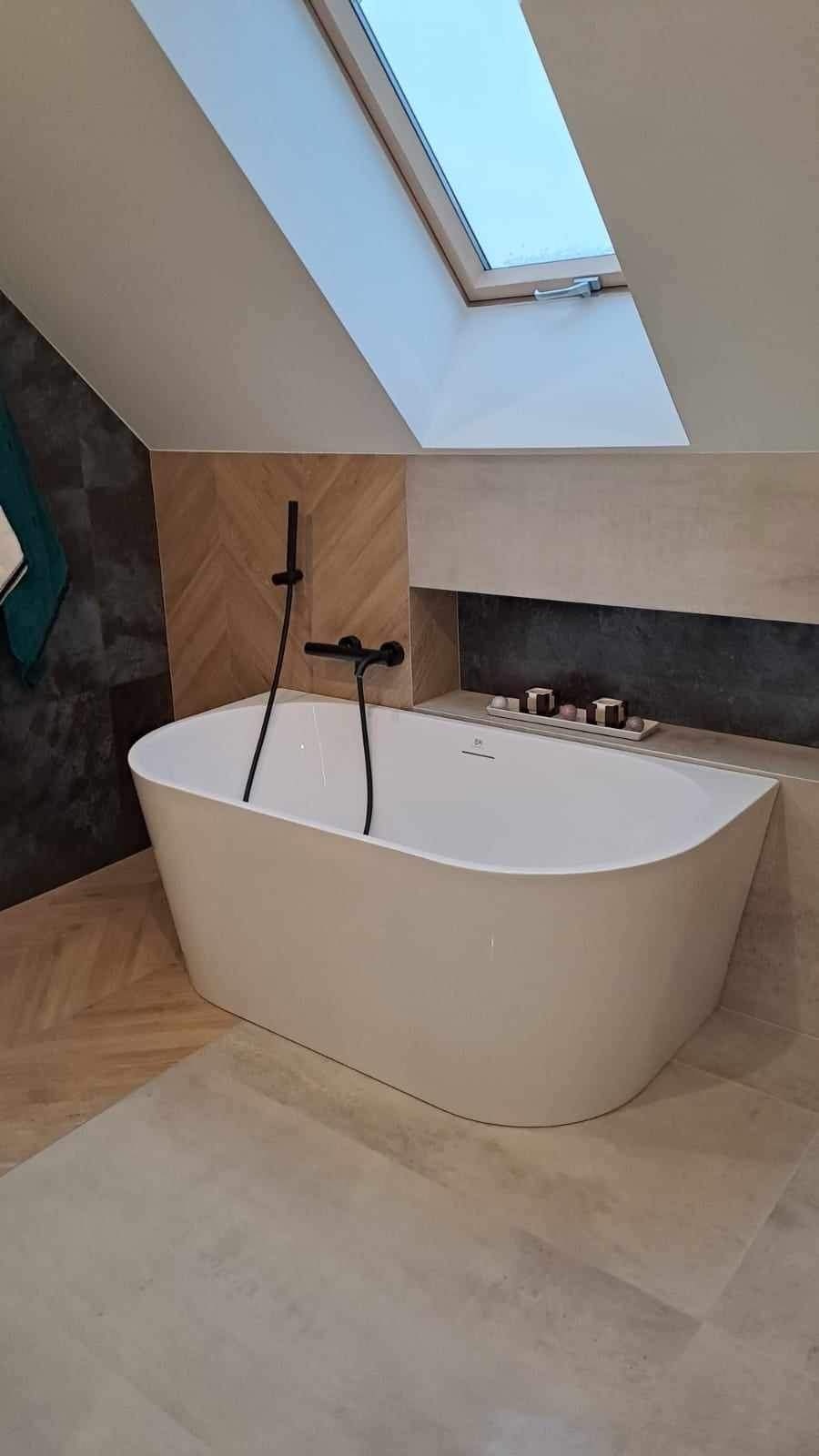 Biała wolnostojąca wanna z czarną armaturą w wykończonej łazience z oknem dachowym, widoczna półka z dekoracjami i ściana z drewnopodobną mozaiką.