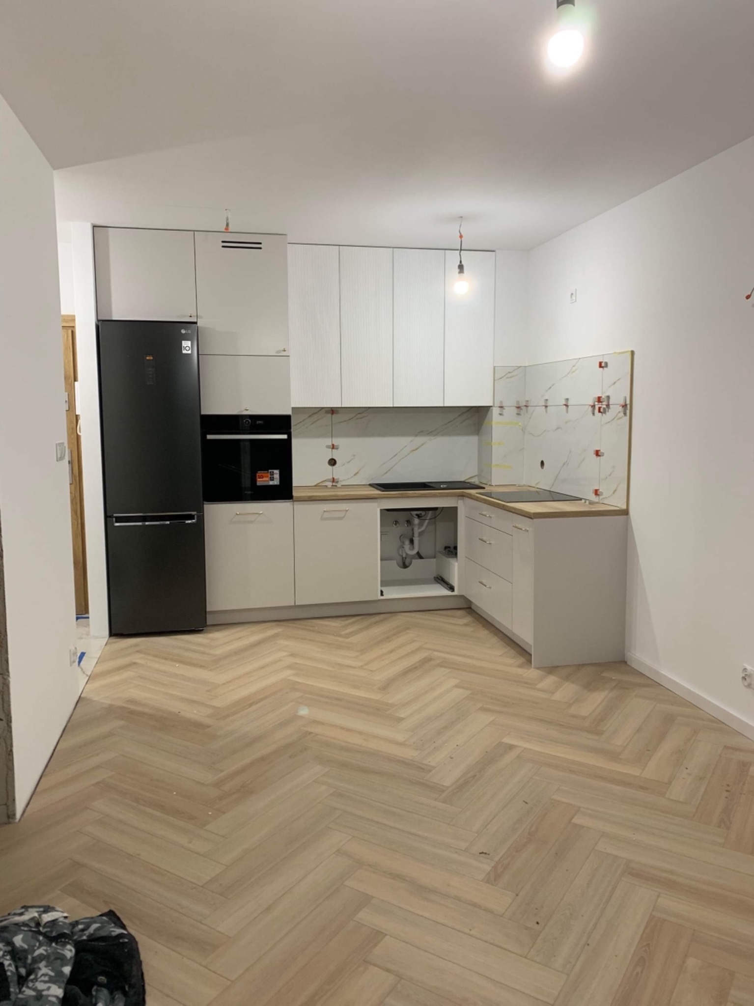 Nowoczesna kuchnia w trakcie wykończenia: białe szafki, czarna lodówka LG, drewniany blat, płytki z marmurowym wzorem, parkiet ułożony w jodełkę, widoczne elementy instalacji hydraulicznej pod zlewem.