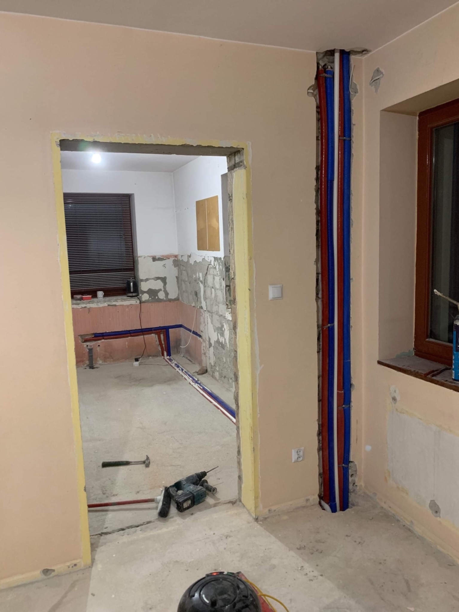 Wnętrze w trakcie remontu: widoczne nowe instalacje hydrauliczne w ścianach, narzędzia na podłodze, ściany w stanie surowym, okno z drewnianą ramą.