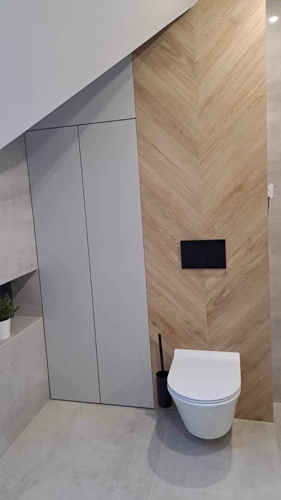 Nowoczesna toaleta podwieszana w łazience na poddaszu, z drewnopodobną ścianą ułożoną w jodełkę i szarą zabudową meblową.