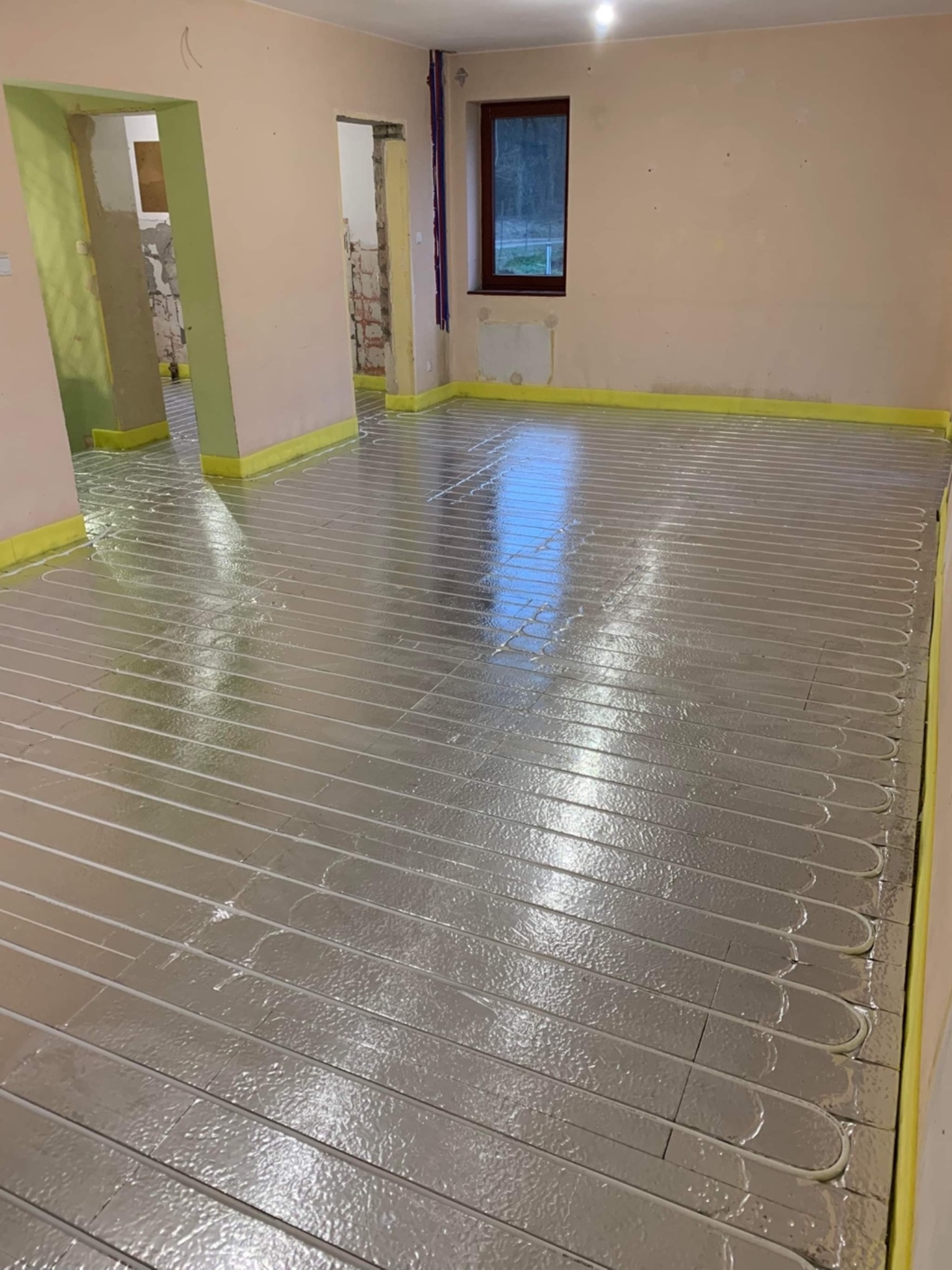 Rozłożone rury ogrzewania podłogowego na folii izolacyjnej, widoczne przygotowania do wylewki, żółta taśma brzegowa przy ścianach.