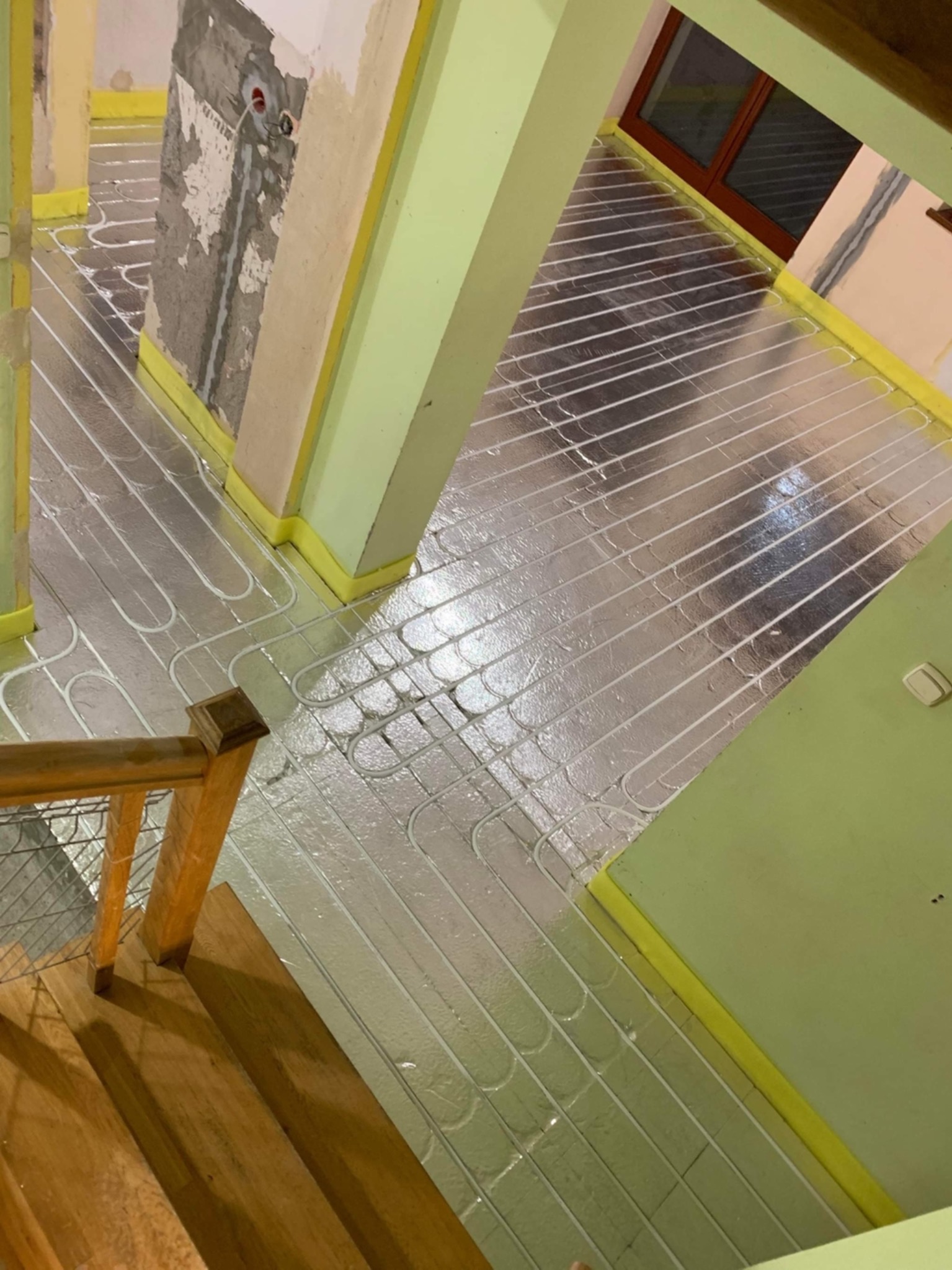 Instalacja ogrzewania podłogowego w trakcie realizacji, widoczne rury grzewcze na folii izolacyjnej, żółta taśma przy ścianach, surowy stan ścian i drewniane schody w tle.