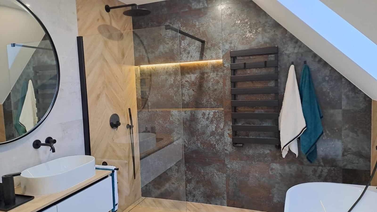 Nowoczesna łazienka na poddaszu z kabiną prysznicową typu walk-in, umywalką nablatową na szafce z białymi frontami, okrągłym lustrem w czarnej ramie i grzejnikiem drabinkowym w kolorze antracytowym.