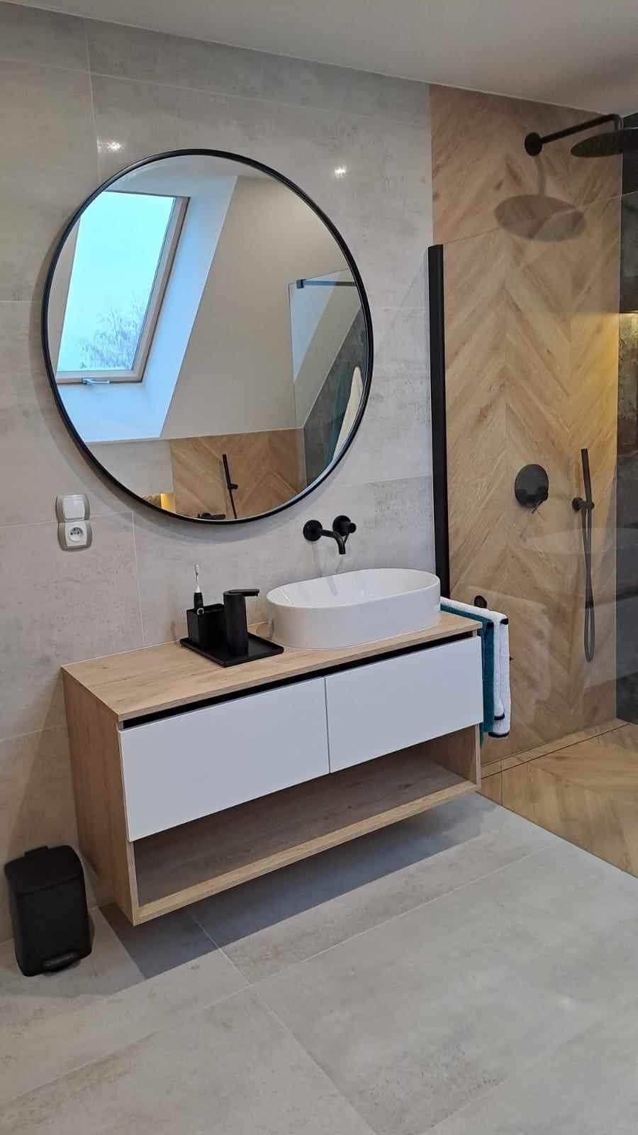 Nowoczesna łazienka z owalną umywalką nablatową, okrągłym lustrem, czarną armaturą i kabiną prysznicową z drewnopodobnymi płytkami ułożonymi w jodełkę.