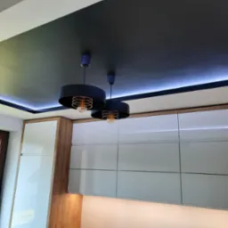 Nowoczesna kuchnia z czarnym sufitem podwieszanym, oświetleniem LED i białymi szafkami z drewnianymi akcentami oraz dwoma okrągłymi lampami wiszącymi z ozdobnymi żarówkami.