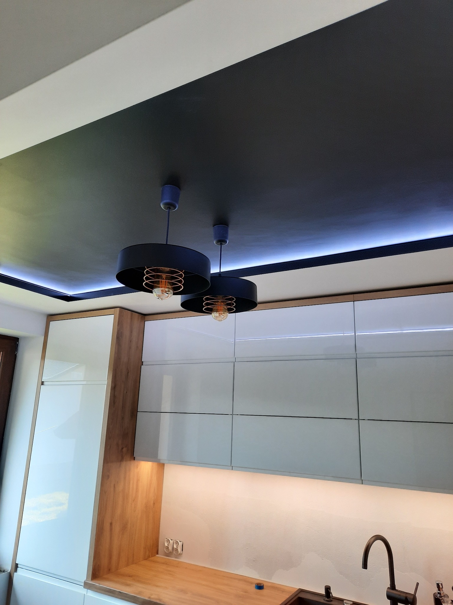 Nowoczesna kuchnia z czarnym sufitem podwieszanym, oświetleniem LED i białymi szafkami z drewnianymi akcentami oraz dwoma okrągłymi lampami wiszącymi z ozdobnymi żarówkami.